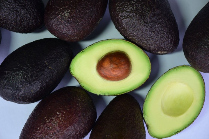Aguacates Hass enteros de piel oscura junto a un aguacate cortado a la mitad mostrando pulpa verde fresca y hueso central.