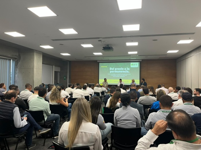 Una gran audiencia presta atención mientras un presentador en el escenario muestra datos técnicos en pantallas.